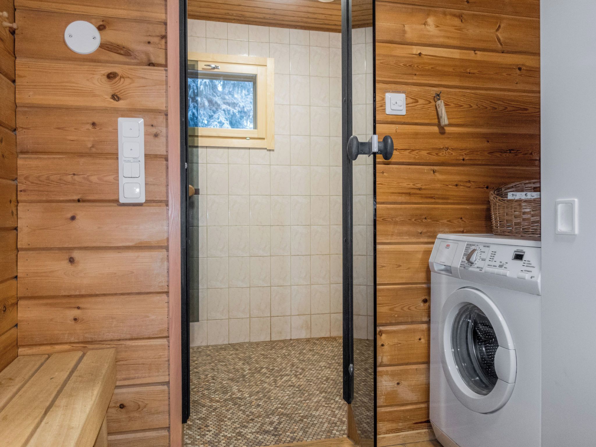 A laundry room with a washing machine, a wooden wall, a tiled floor, and a door leading to a shower area.