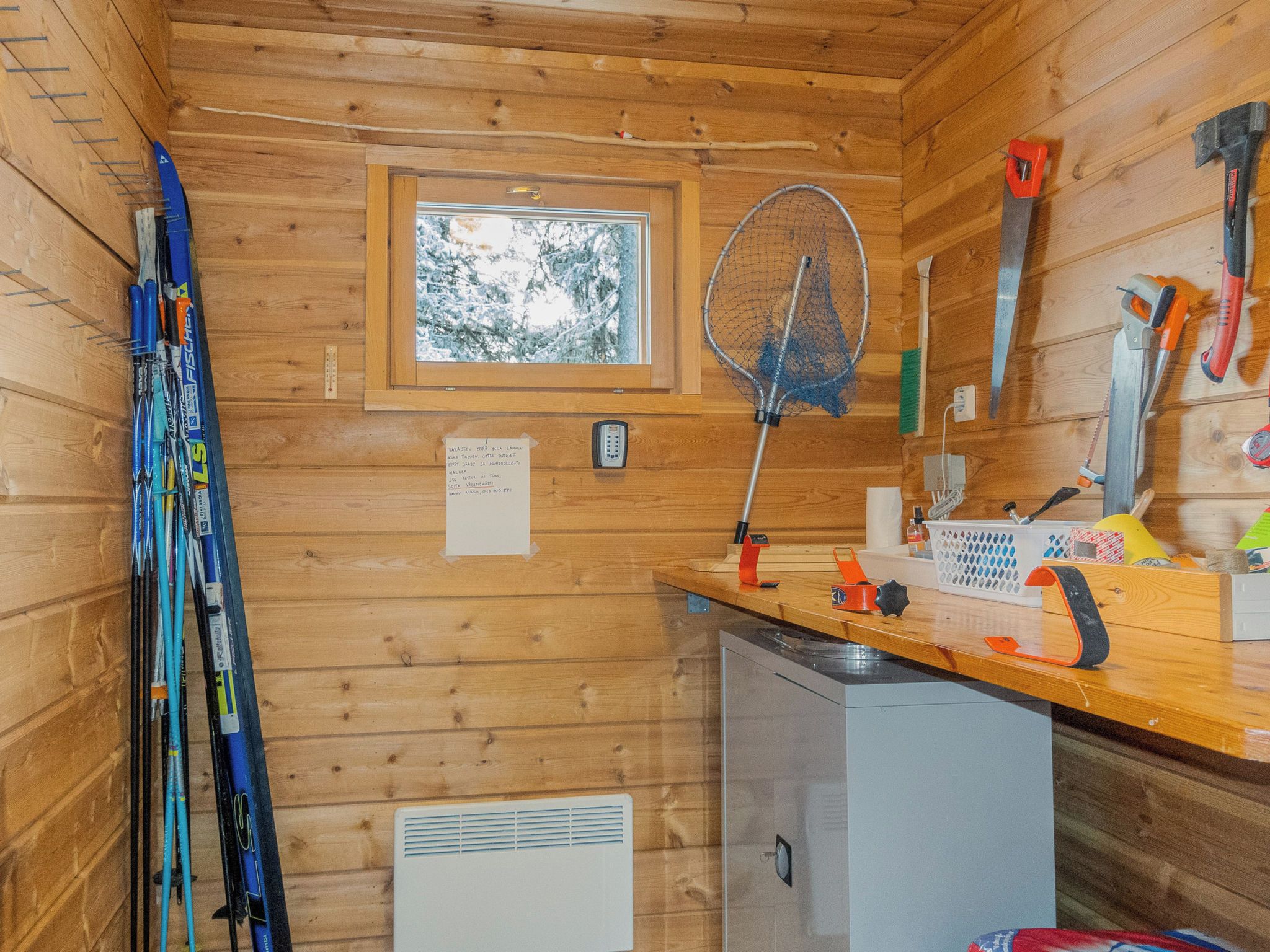 A wooden cabin interior with a window, a small refrigerator, and various tools and equipment.