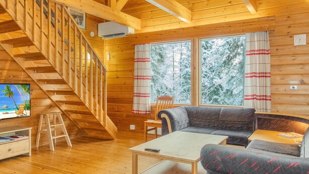 A wooden cabin interior with a staircase, a sofa, a table, and a TV on a wooden stand.