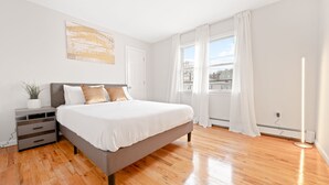 Een moderne slaapkamer met een groot bed, een nachtkastje met lades, een kamerplant en een uitzicht buiten door een raam met witte gordijnen.