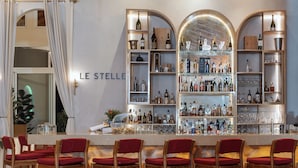 Een bar met meerdere flessen en glazen op planken, houten stoelen met rode kussens, en een bord waarop 'LE STELLE' staat.