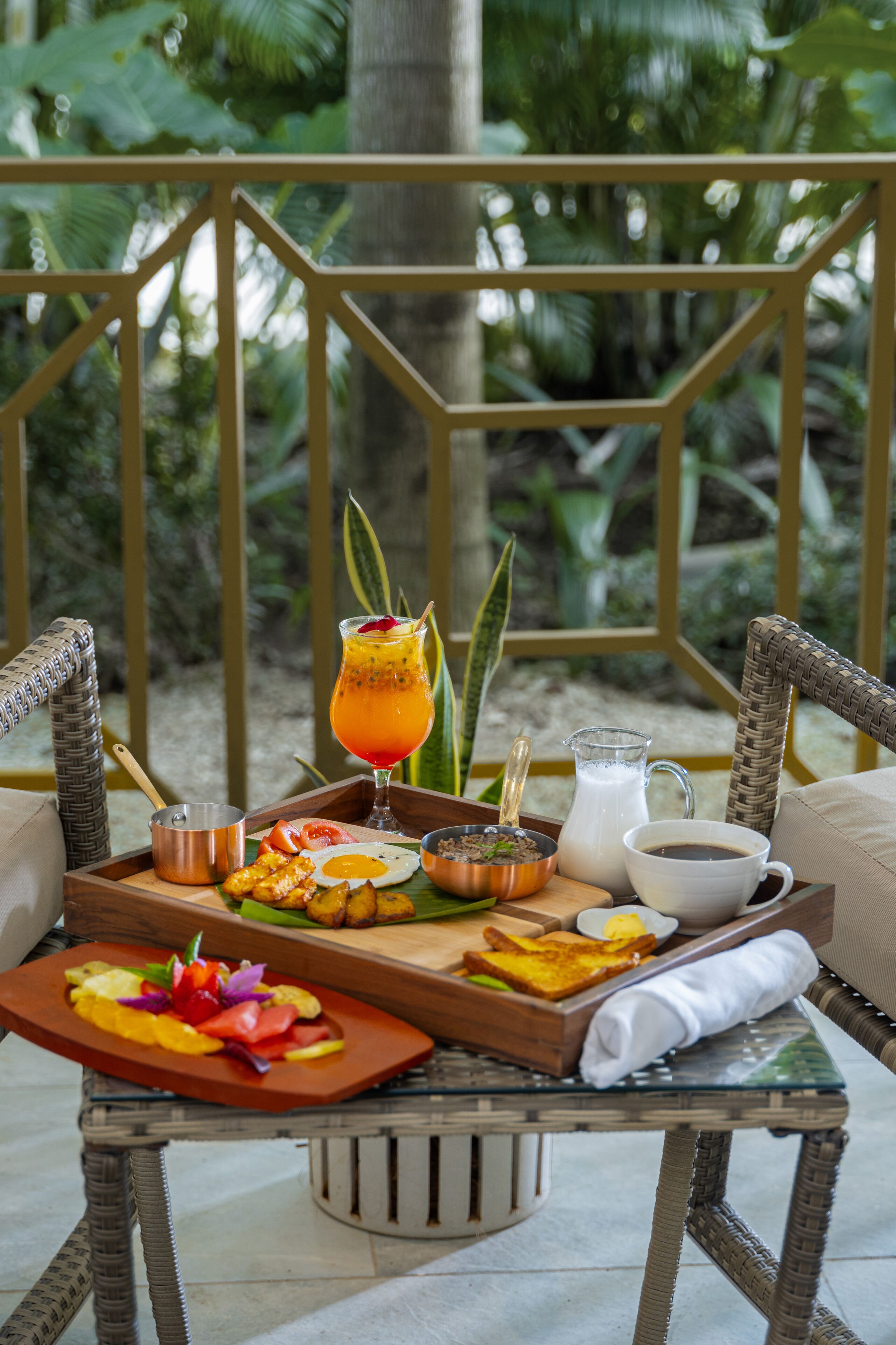 Un plateau de petit-déjeuner contenant divers aliments et boissons, posé sur une table à l’extérieur, entouré de chaises en osier et d’une rambarde.