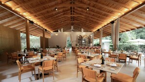 Een ruim restaurant met houten tafels en stoelen, grote ramen en een hoge plafond.