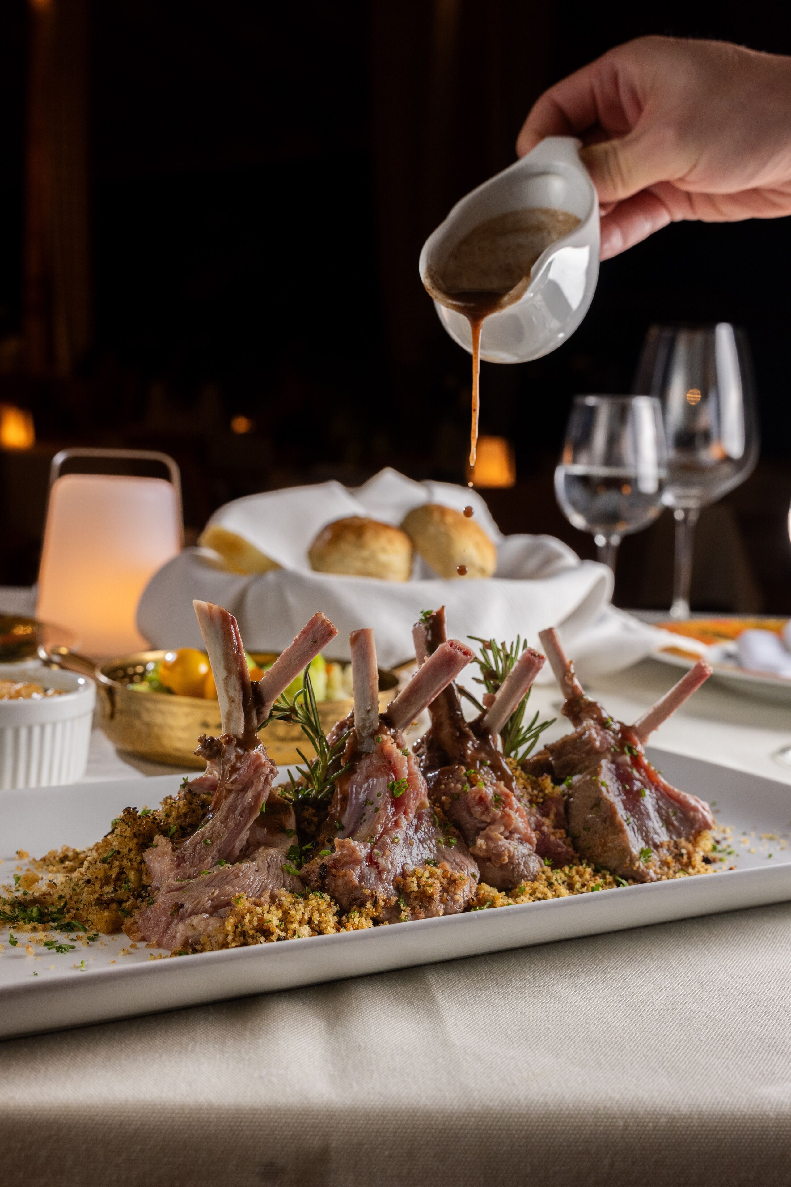 A dish of lamb chops with a sauce being poured over them, accompanied by bread and a candle in the background.