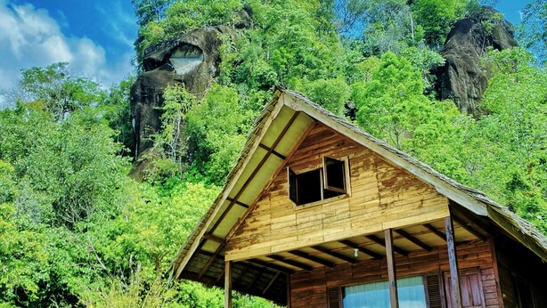 A wooden cabin with a porch, surrounded by lush greenery and rocky cliffs.