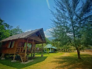 A rustic wooden cabin with a thatched roof, a covered porch, and a small garden area. - Rickys Beach House (Koto XI Tarusan)