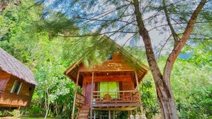 A wooden cabin with a thatched roof, surrounded by lush greenery and tall trees.