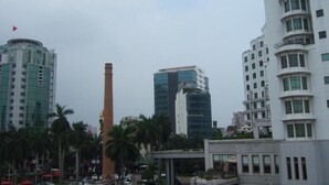 A cityscape with modern buildings, including a tall structure with a flag, a cylindrical tower, and a multi-story building with balconies. - Lao Cai Hotel by Bay Luxury (Hanoi)