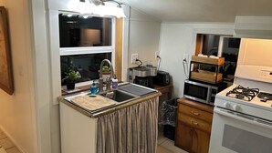 Eine kleine Küche mit Spüle, Herd und Mikrowelle. Über der Spüle befindet sich ein Fenster, und es gibt eine kleine Arbeitsfläche mit Schneidebrett und Seifenspender.