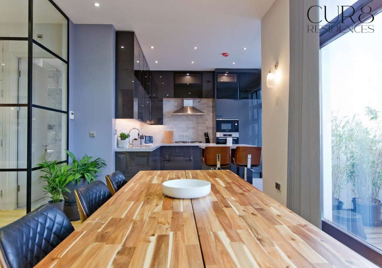A modern kitchen with dark cabinetry, a central island, and a dining area with a wooden table and leather chairs.