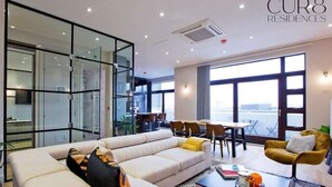 Uma sala de estar moderna com sofá, mesa de centro e cadeiras. Há uma divisória de vidro e vista para a cidade.
