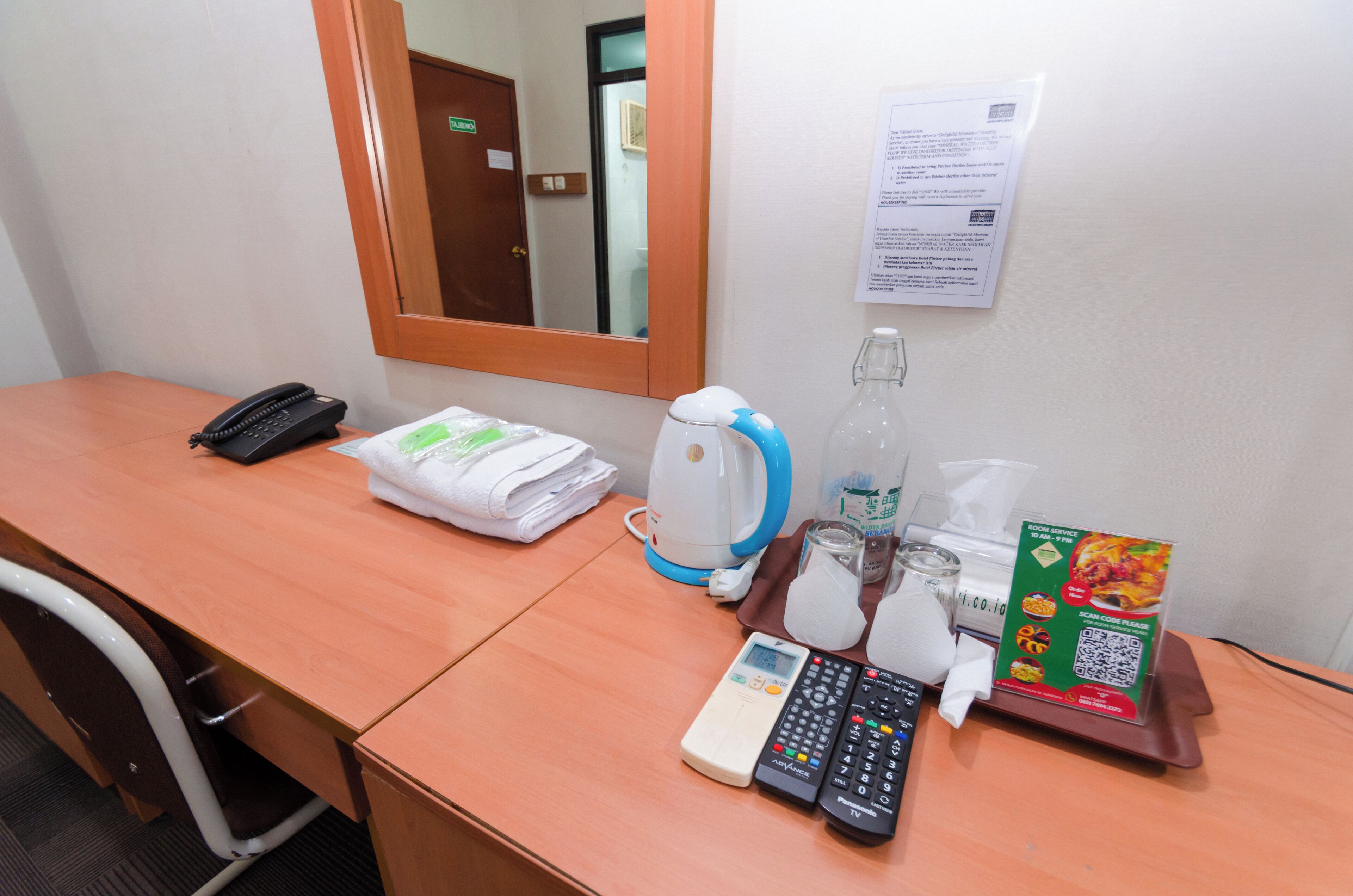 Mesa de quarto de hotel com telefone, chaleira, garrafa, copo, controle remoto e cardápio.