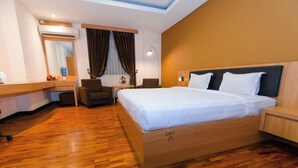 A hotel room with a wooden floor, a bed with white linens, a desk with a computer, a chair, a lamp, and a large mirror. - Graha Widya Bhakti at STIESIA Surabaya (Surabaya)