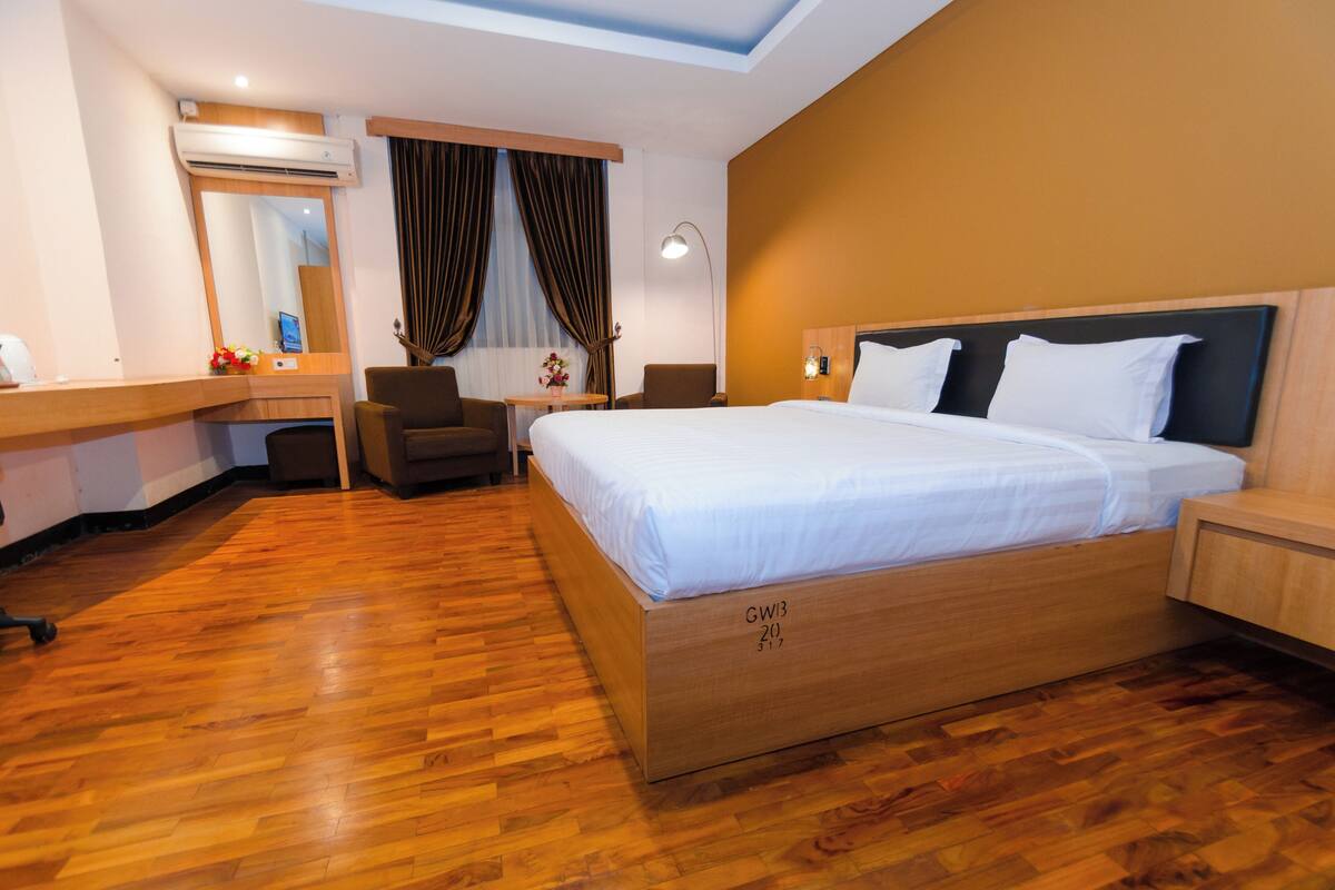 Une chambre d’hôtel avec un plancher en bois, un lit avec du linge de maison blanc, un bureau avec un ordinateur, une chaise, une lampe et un grand miroir.