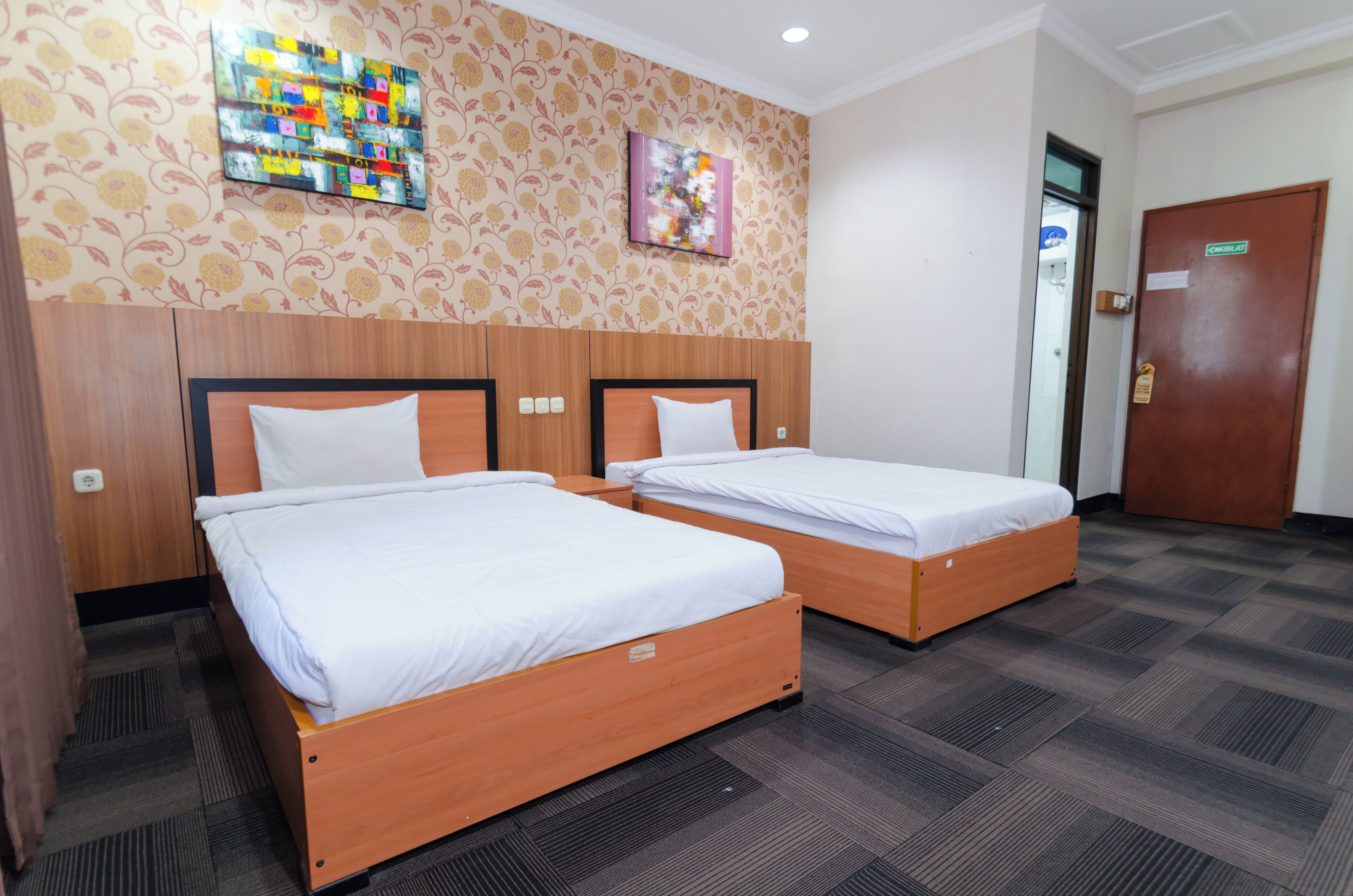 A hotel room with two single beds, a wooden headboard, patterned wallpaper, and a carpeted floor.