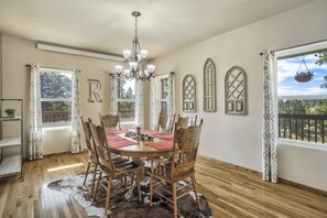 A dining room with a wooden table, chairs, and a chandelier. There are windows with curtains and decorative wall art.