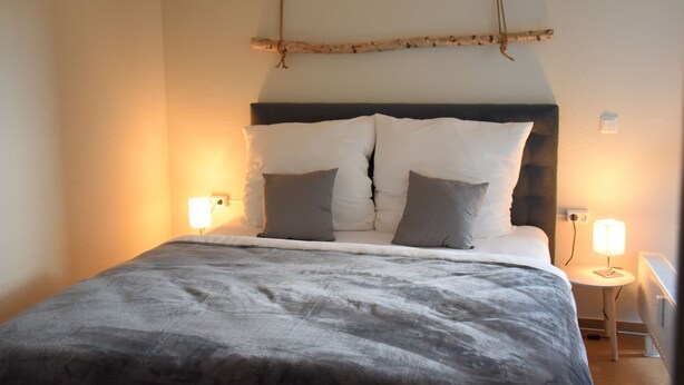 A neatly made bed with a grey blanket, two pillows, and a wooden branch decoration above it. There are two bedside lamps on either side of the bed. - Charming Homes - Studio 25 (Weyhausen)