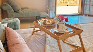 Un dormitorio con una cama, una mesita de noche con una bandeja de pasteles, una taza y vistas a la montaña desde una puerta abierta.