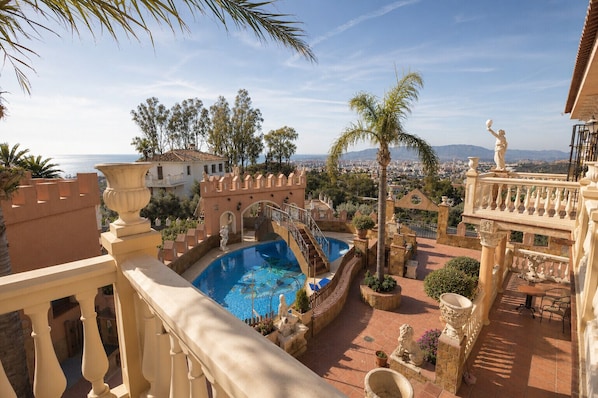 Ein Pool mit einer Steinbrücke, Palmen und Blick auf die Stadt.