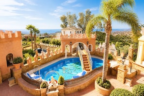 Ein Schwimmbecken mit einer schlossartigen Struktur, einer Treppe, die zum Pool führt, und Blick auf die Stadt im Hintergrund.