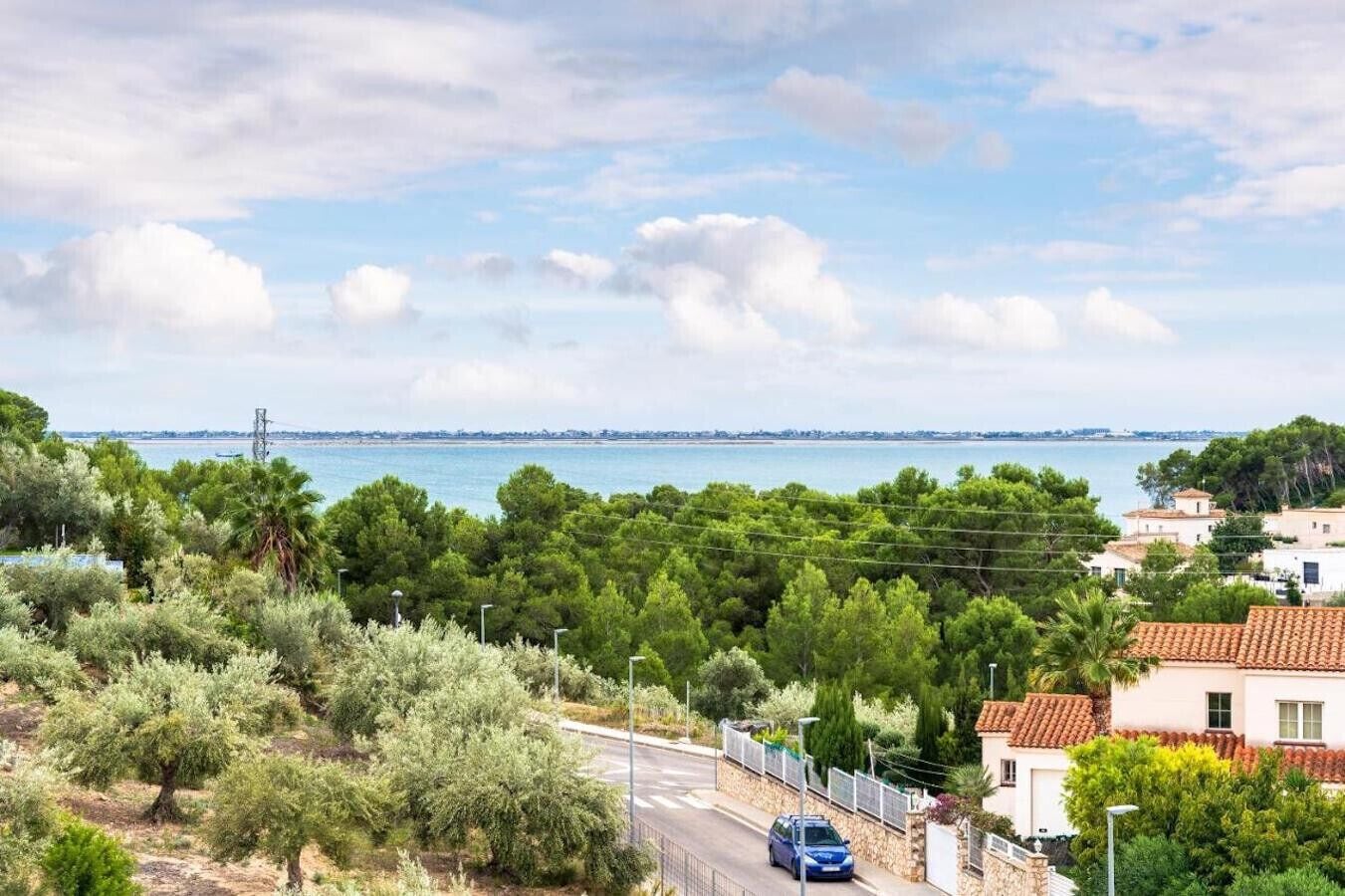 Une route côtière bordée d’une voiture bleue, entourée d’une végétation luxuriante et de maisons aux toits recouverts de tuiles rouges.