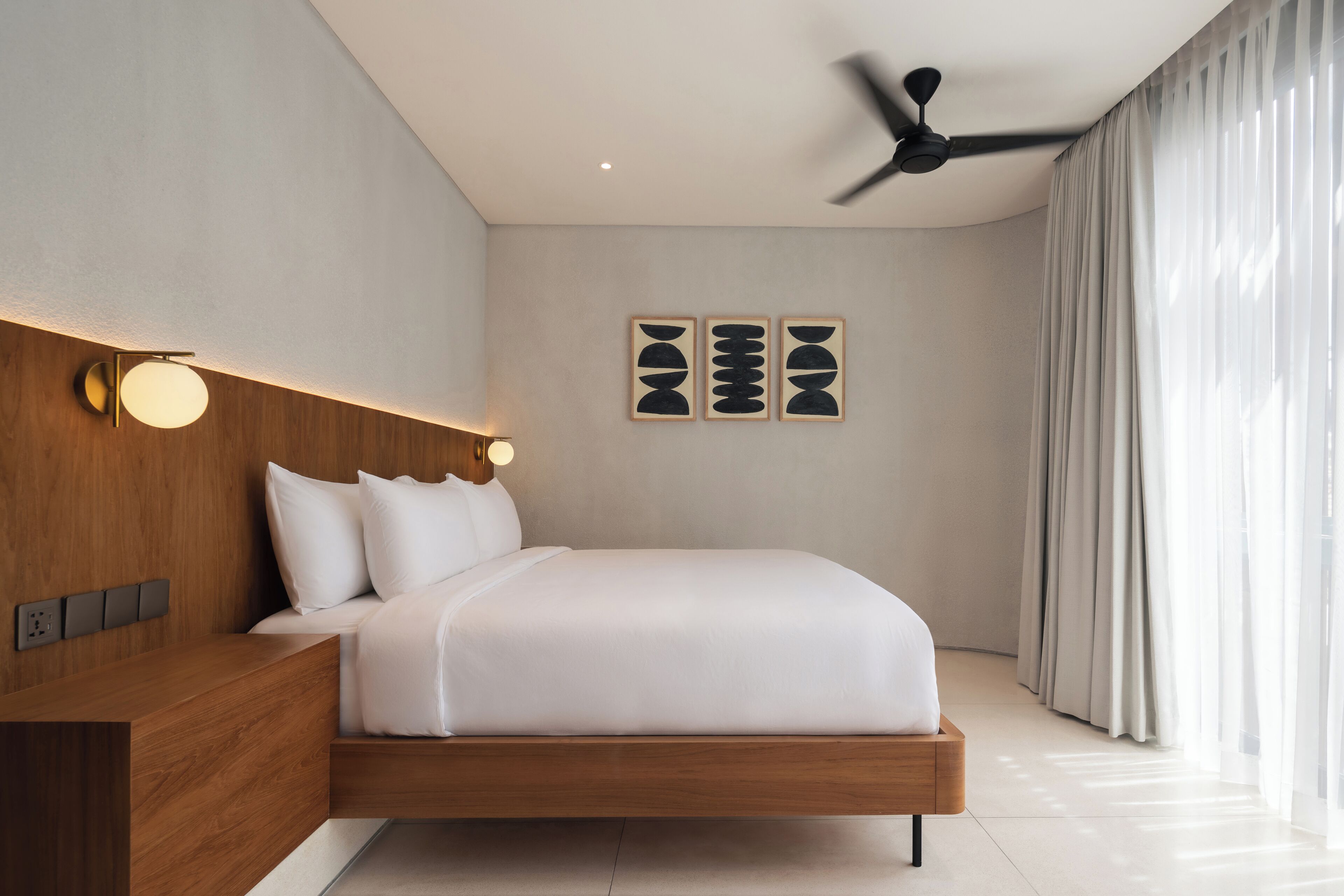 Une chambre d’hôtel moderne avec une tête de lit en bois, un lit avec une literie blanche, un ventilateur de plafond et des œuvres d’art accrochées au mur.