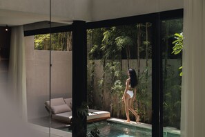 A modern indoor pool area with a glass wall, a sofa, and bamboo plants. - VOX Villas (Canggu)
