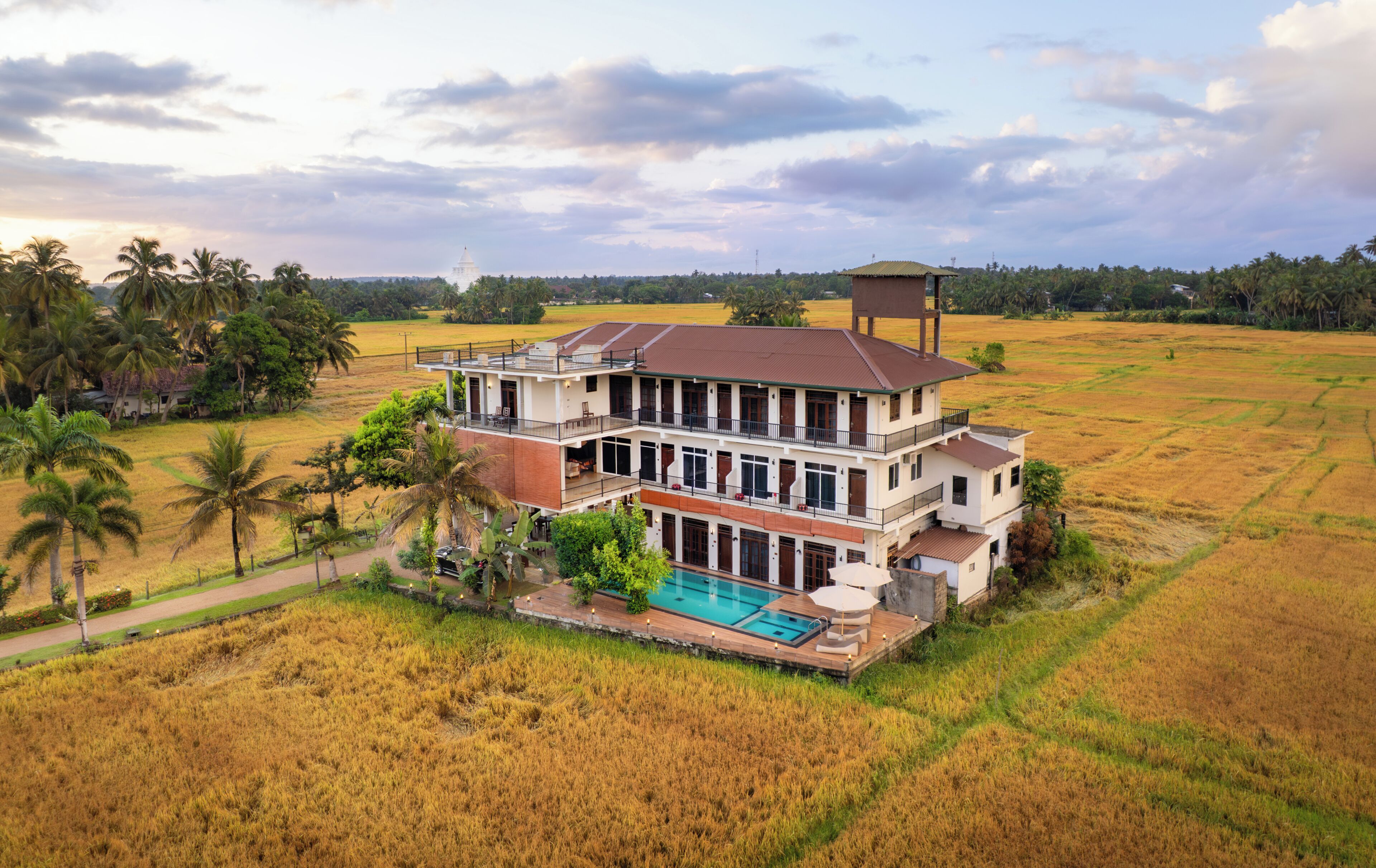 Una amplia casa de dos pisos con piscina, rodeada de campos y palmeras.