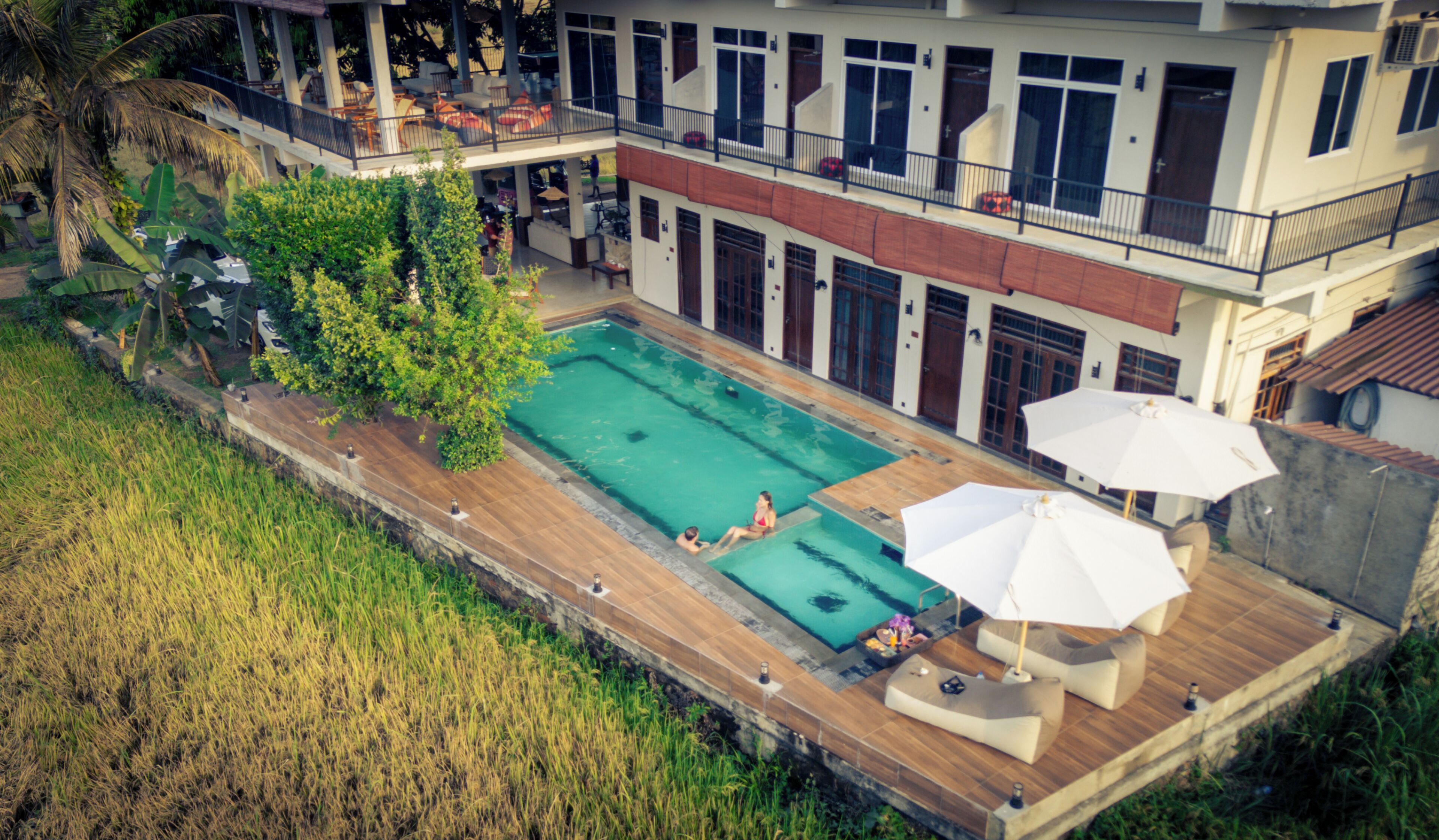 Una amplia piscina al aire libre con terraza de madera, rodeada de abundante vegetación y un edificio de varios pisos con balcones.