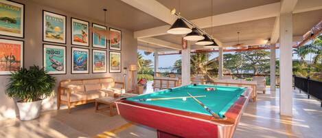 A pool table in a well-lit room with framed pictures on the wall, a sofa, and a potted plant.