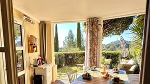 An open patio with a wooden table and chairs, a small kitchen area with a coffee maker, and a view of a lush garden. - The Garden by the Sea (Saint-Raphaël)
