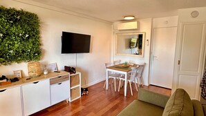 A living room with a green sofa, a dining table with chairs, a TV mounted on the wall, and a small side table with decorative items. - The Garden by the Sea (Saint-Raphaël)