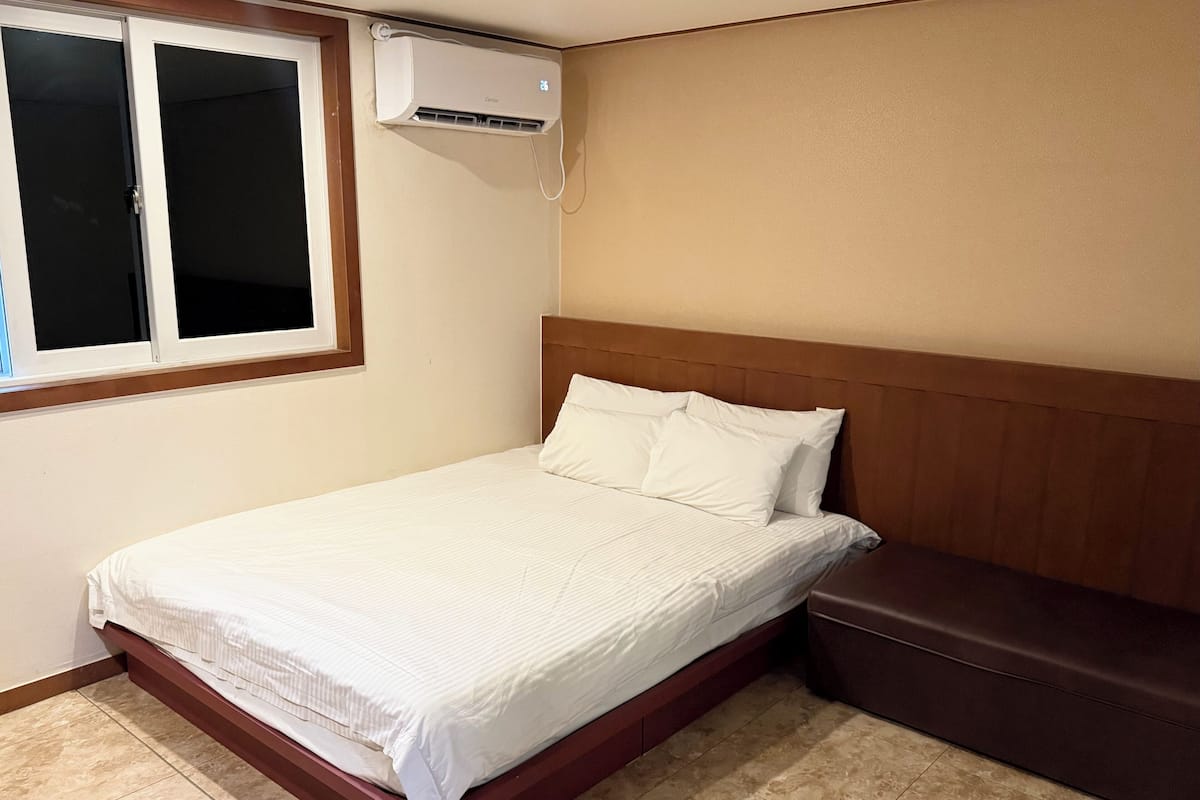 A single bed with a wooden headboard, a window with blinds, and an air conditioning unit mounted on the wall.