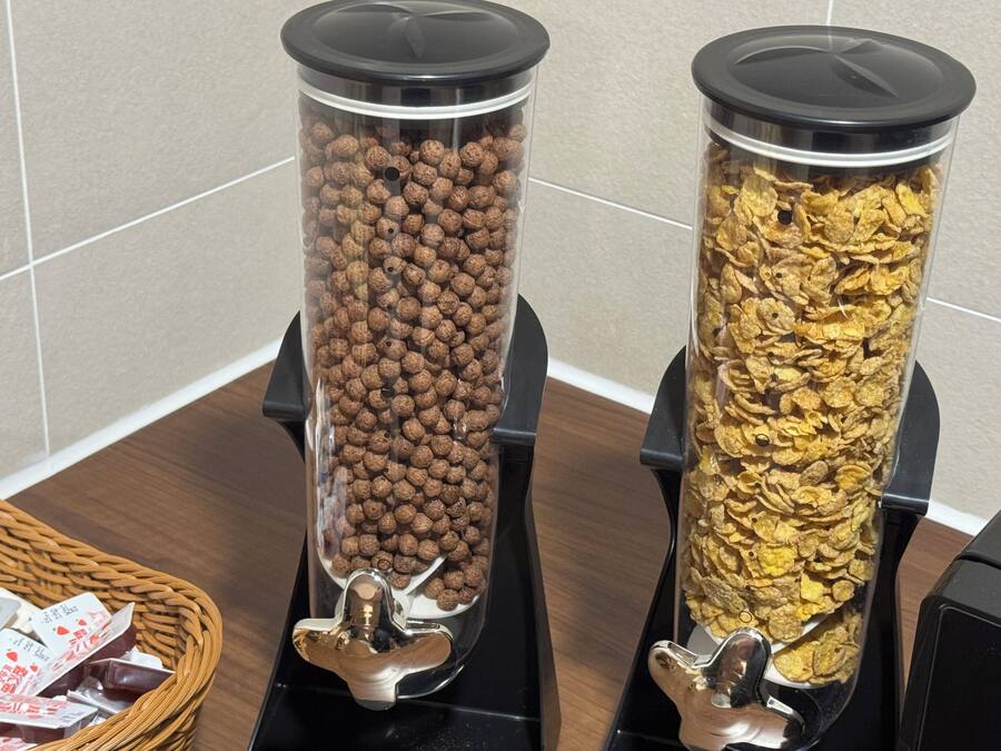 Deux distributeurs cylindriques remplis d’en-cas sont placés sur un comptoir : l’un contient des boules de chocolat et l’autre, des céréales en flocons. Un mur carrelé se trouve en arrière-plan.