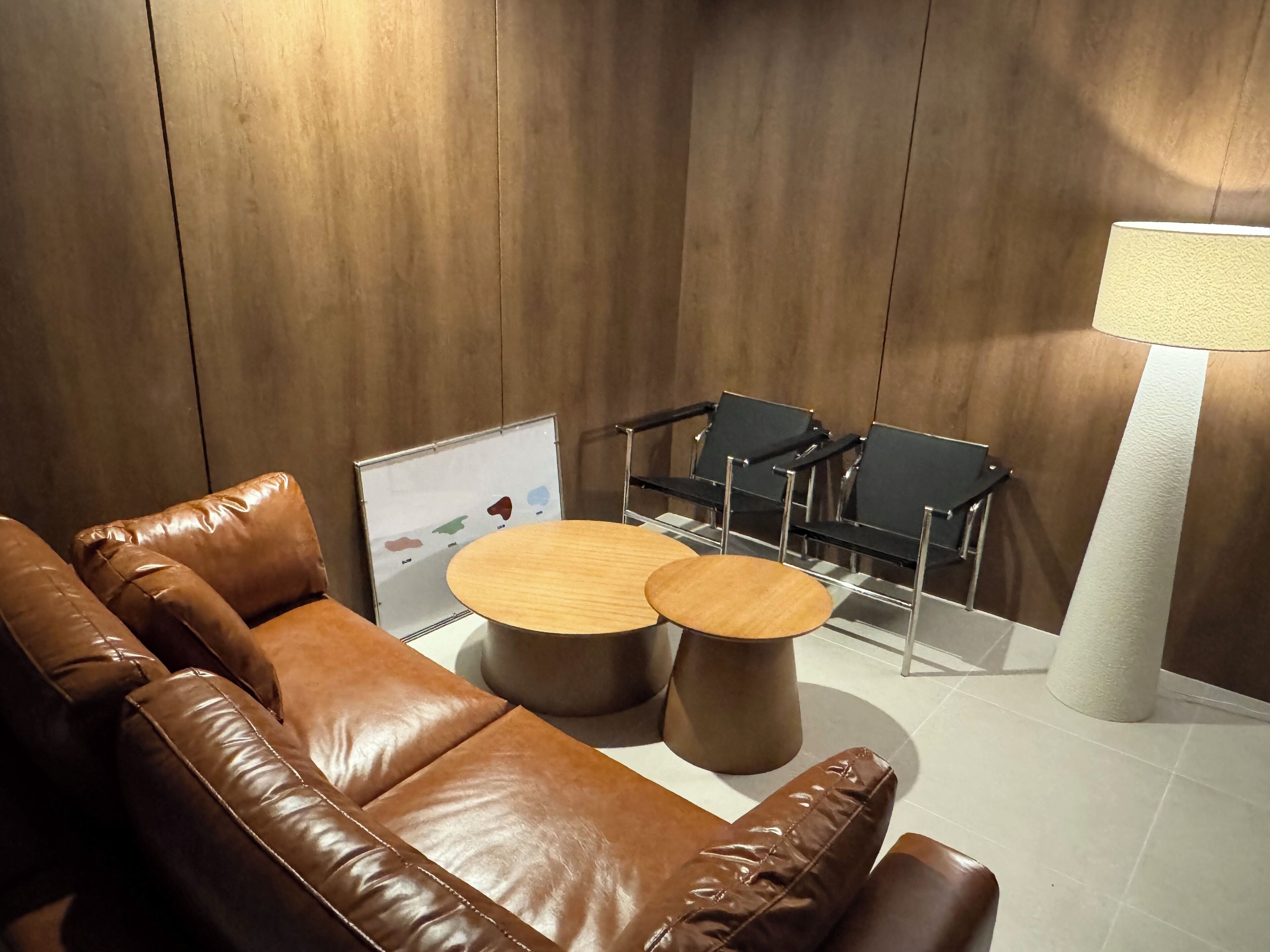 A modern lounge area with a brown leather sofa, two round wooden tables, black chairs, and a white lamp.