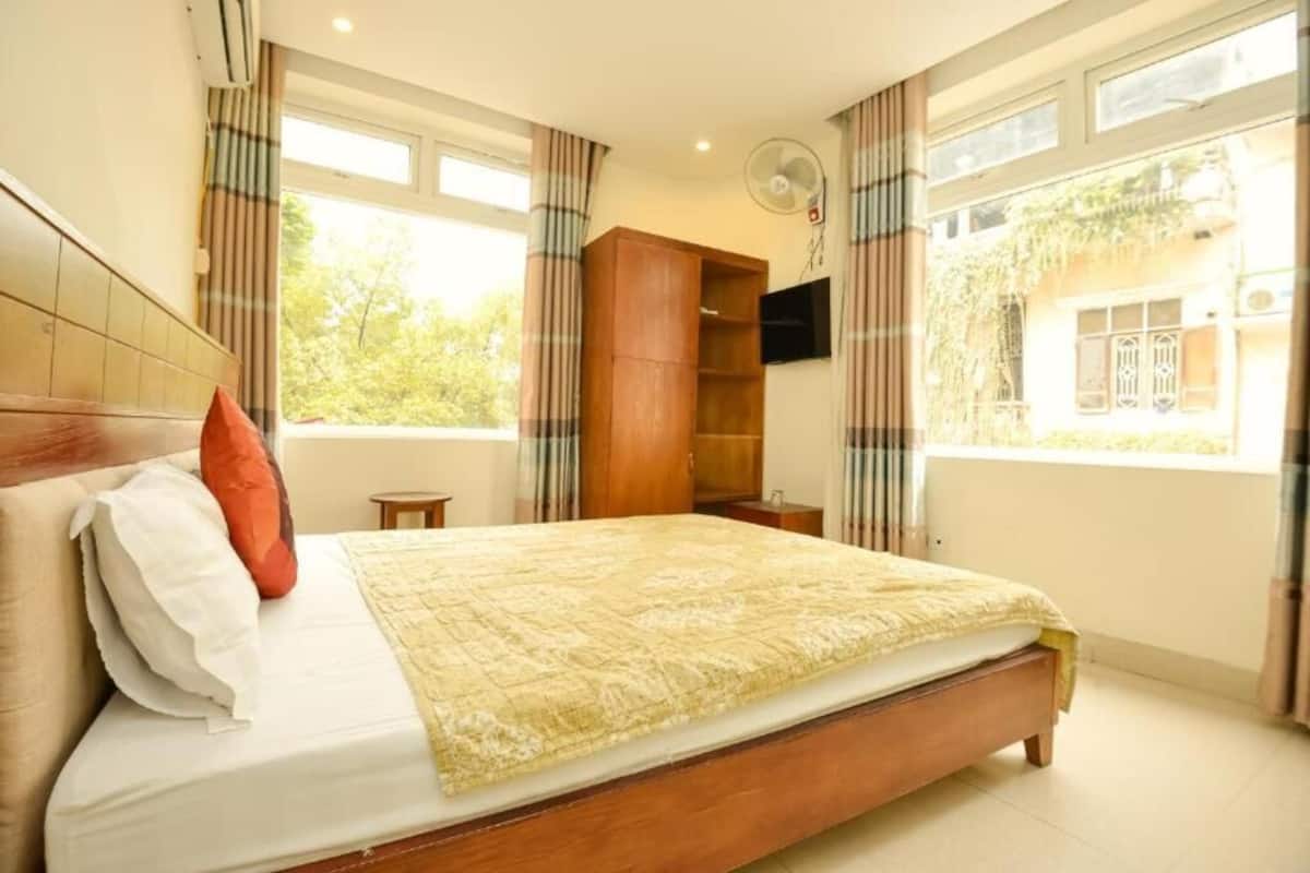 Une chambre à coucher avec un lit, une armoire en bois, un téléviseur fixé au mur et une fenêtre avec des rideaux.
