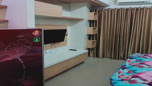A bedroom with a TV mounted on the wall, a bed with a floral bedspread, a refrigerator, and a window with curtains. - OYO 93543 Belmount Residence Near SILOAM CLINIC MERUYA (Jakarta)