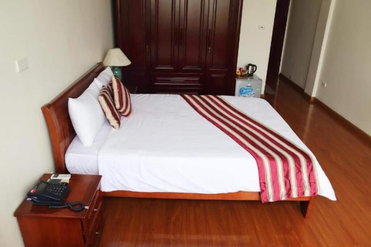 A neatly made bed with white bedding and a red and white striped blanket, a wooden nightstand with a telephone, and a wooden wardrobe in the background.