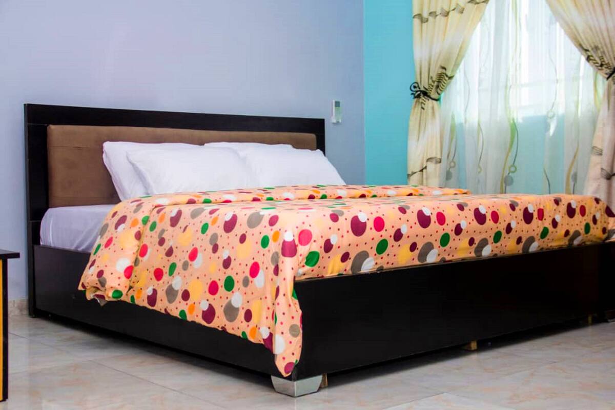 A bed with a polka dot cover, a wooden headboard, and a window with sheer curtains.