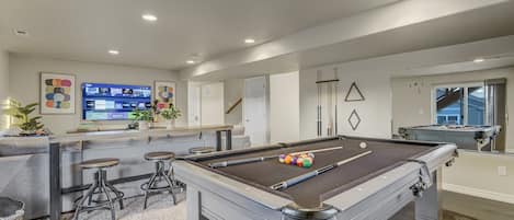 A pool table with stools, a bar area with a TV, and a pool table in a modern room.