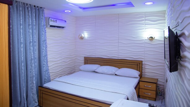 A bedroom with a wooden bed, a nightstand, a TV mounted on the wall, and a ceiling light fixture. - Haffy Executive Hotels and Suite (Uyo)