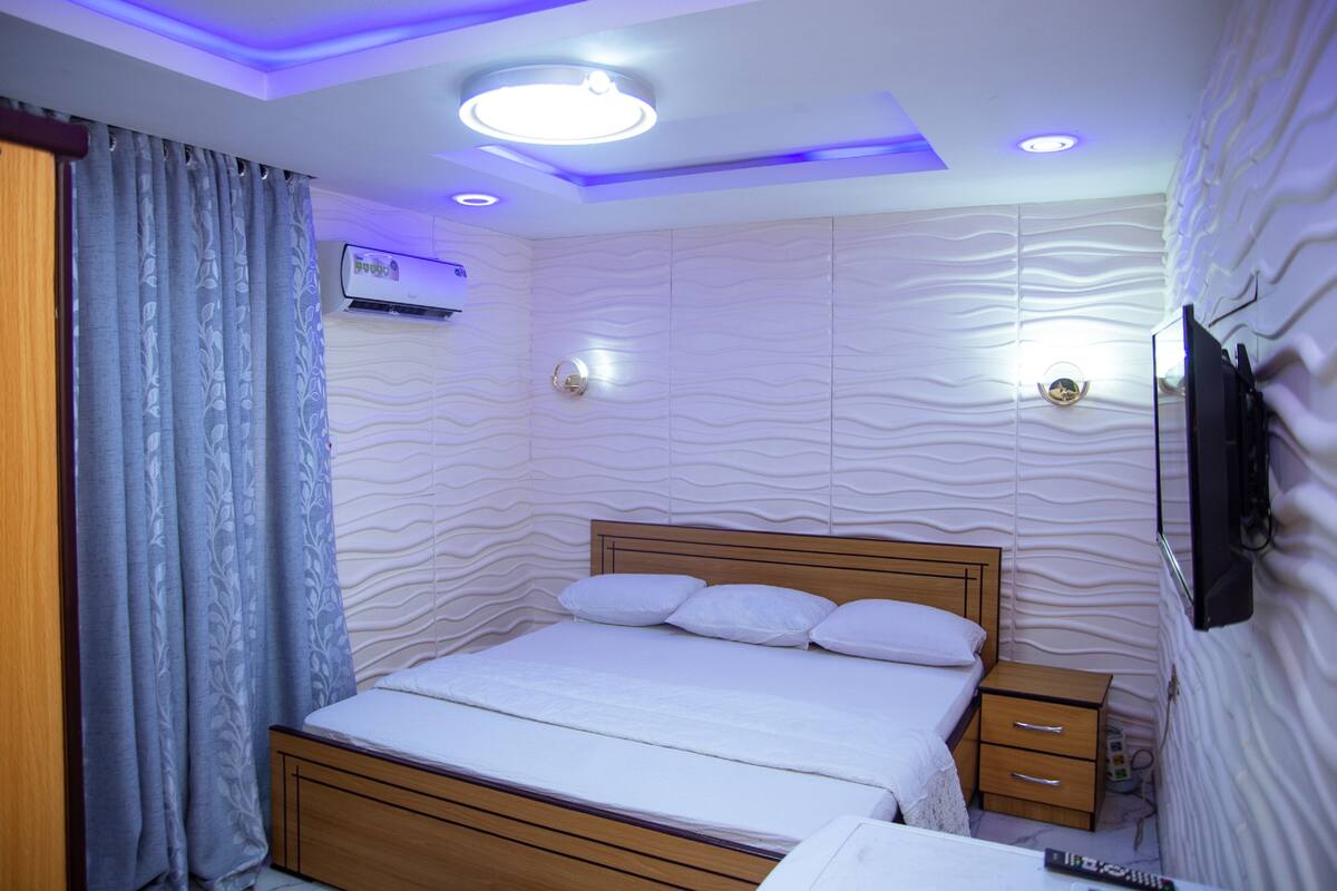 A bedroom with a wooden bed, a nightstand, a TV mounted on the wall, and a ceiling light fixture.