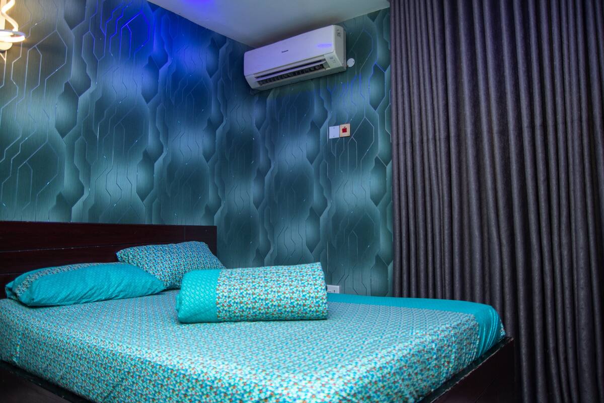 A bedroom with a patterned wall, a bed with teal bedding, an air conditioning unit, and a light switch.