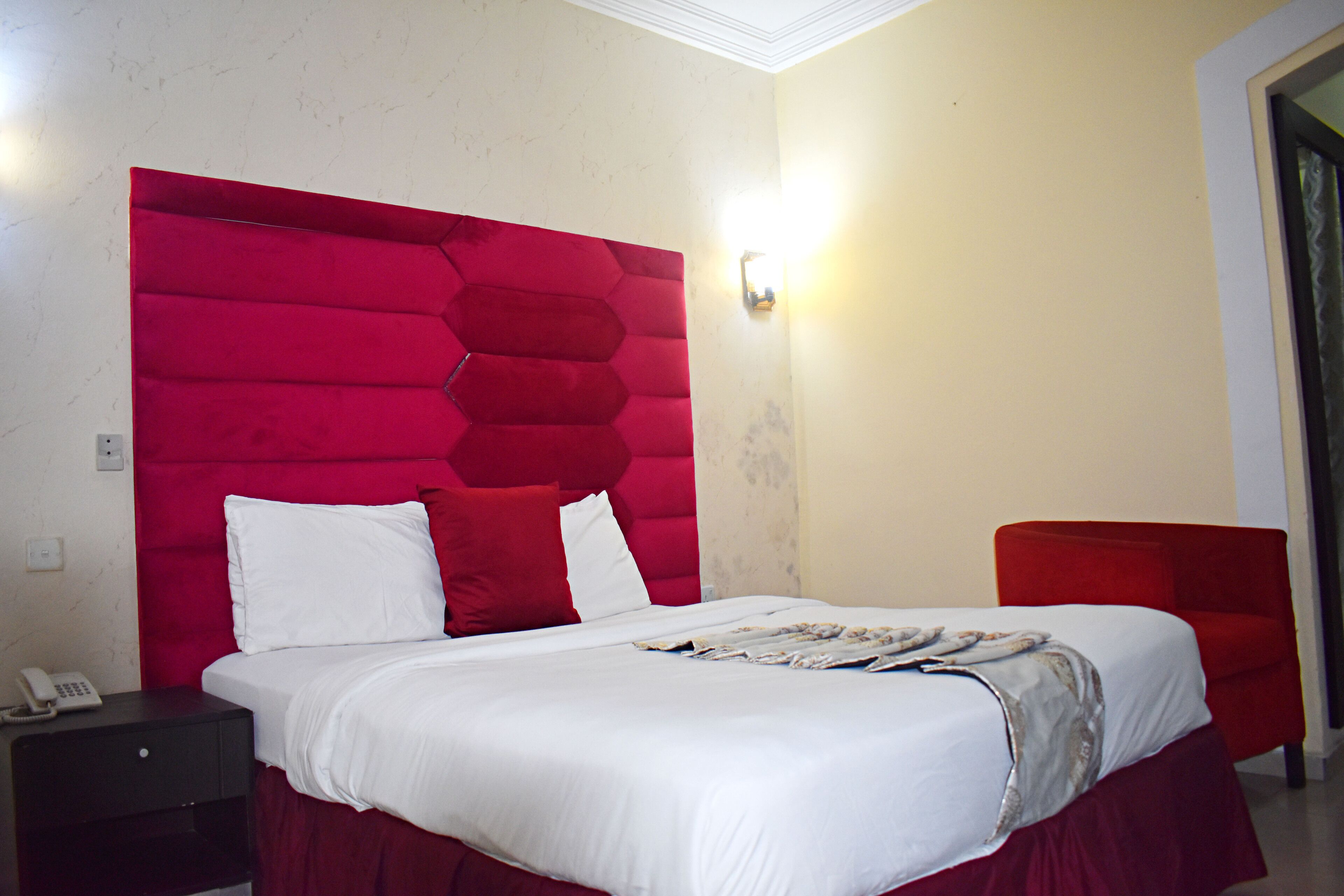 A hotel room with a large bed, a red headboard, a bedside table with a phone, and a red chair.
