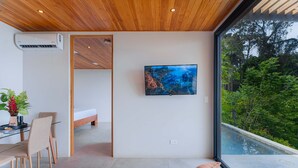 Quarto moderno com cama, televisão, área de refeições e uma janela ampla com vista para vegetação.