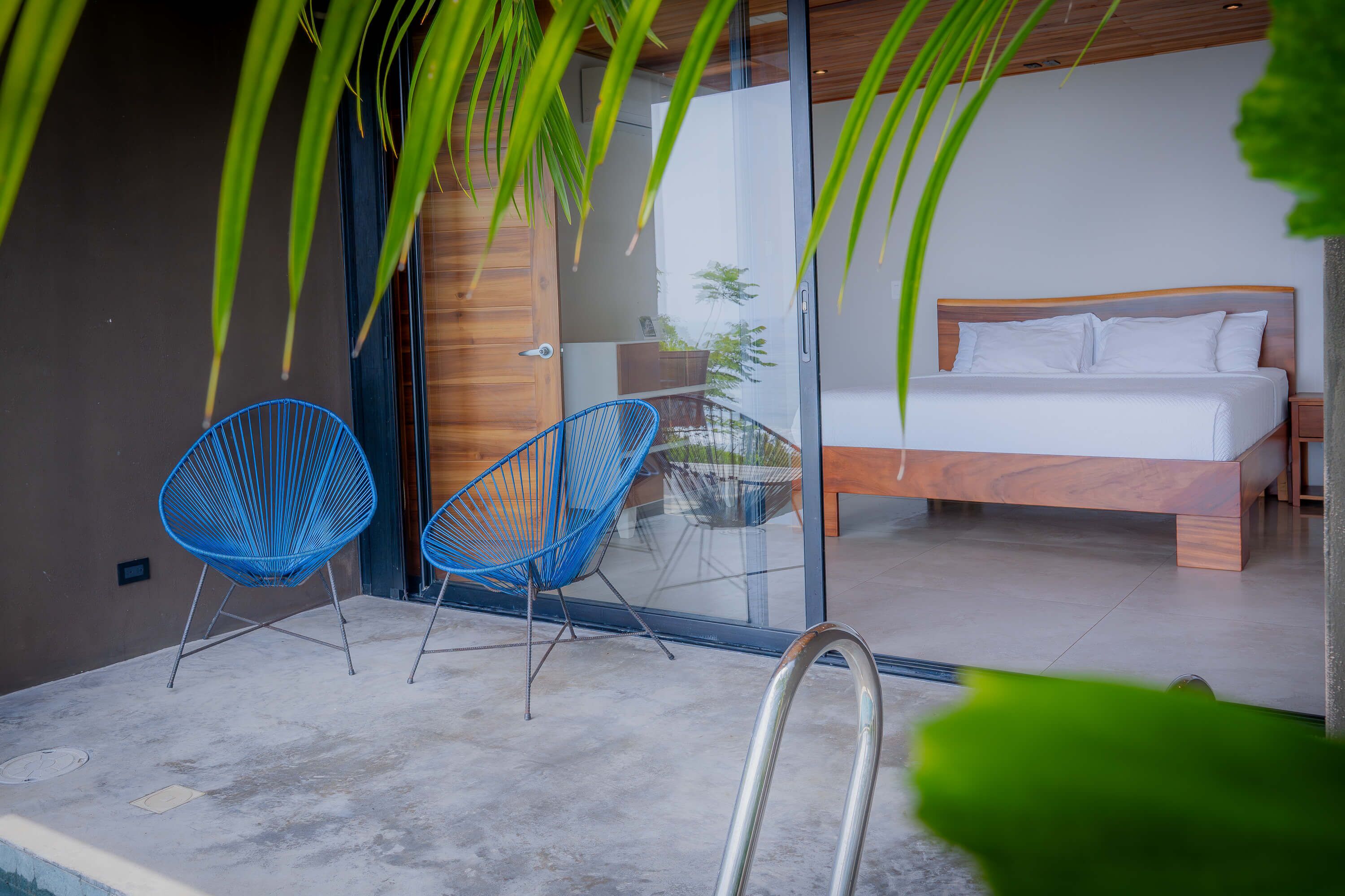 A modern bedroom with a wooden bed, two blue chairs, and a glass door leading to a balcony.