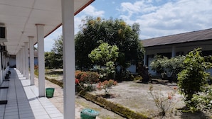 A covered outdoor area with a tiled floor, a green trash bin, and a small garden with various plants and a tree. - Hotel O Pondok Gusang Kuala Near Pelabuhan H Basran (Sampit)