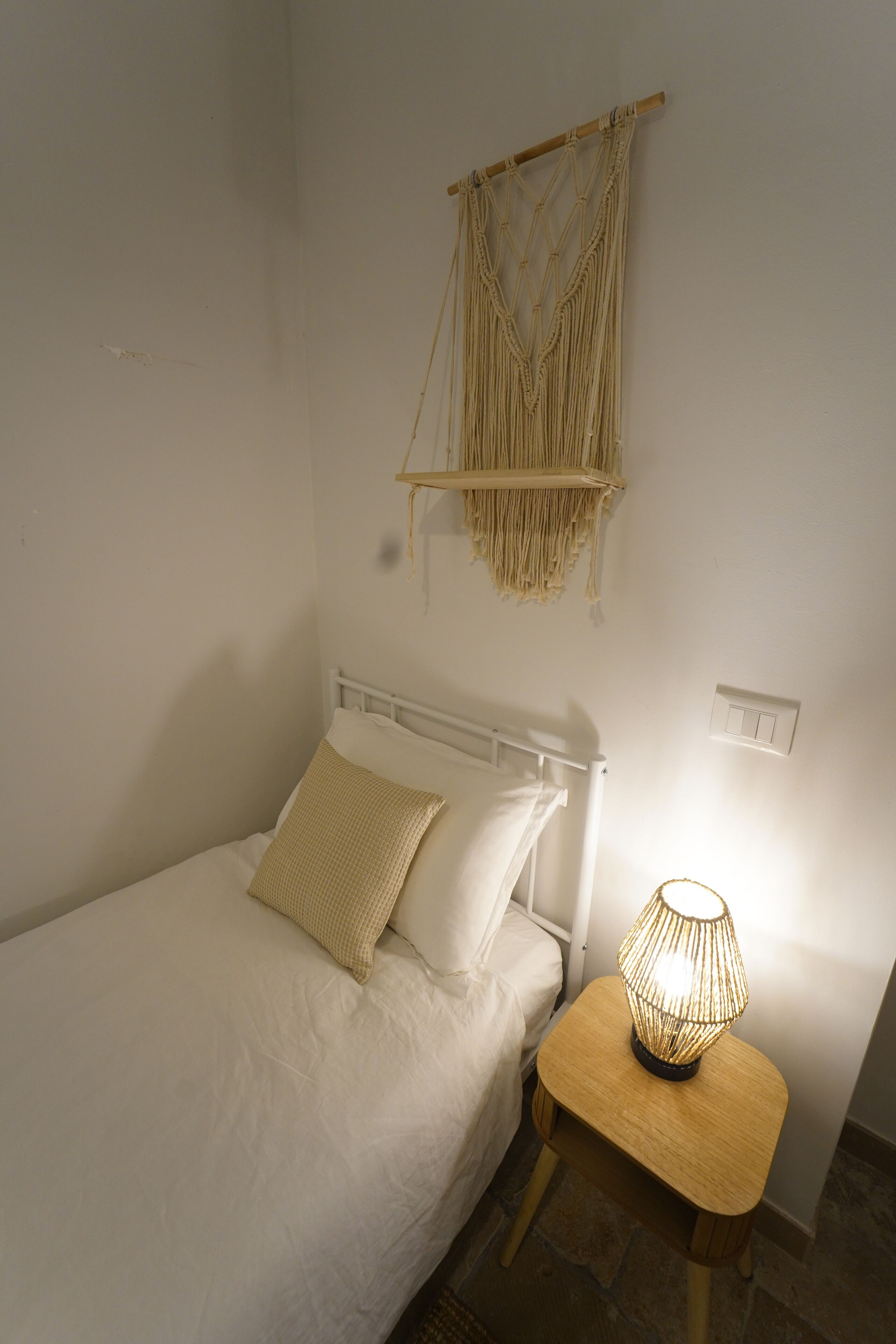 Un lit avec une literie blanche, une table de chevet avec une lampe et une décoration murale en macramé.