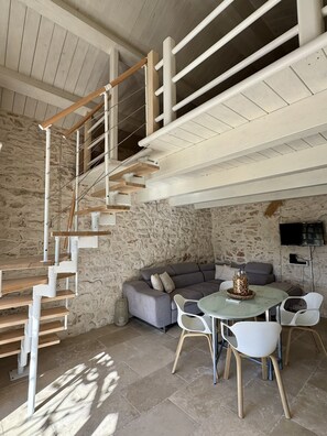 A modern interior with a wooden staircase, stone walls, a gray sofa, and a dining area with white chairs.