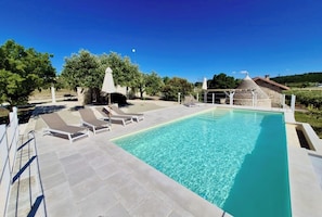 A rectangular swimming pool with clear blue water, surrounded by a tiled deck with lounge chairs and umbrellas.